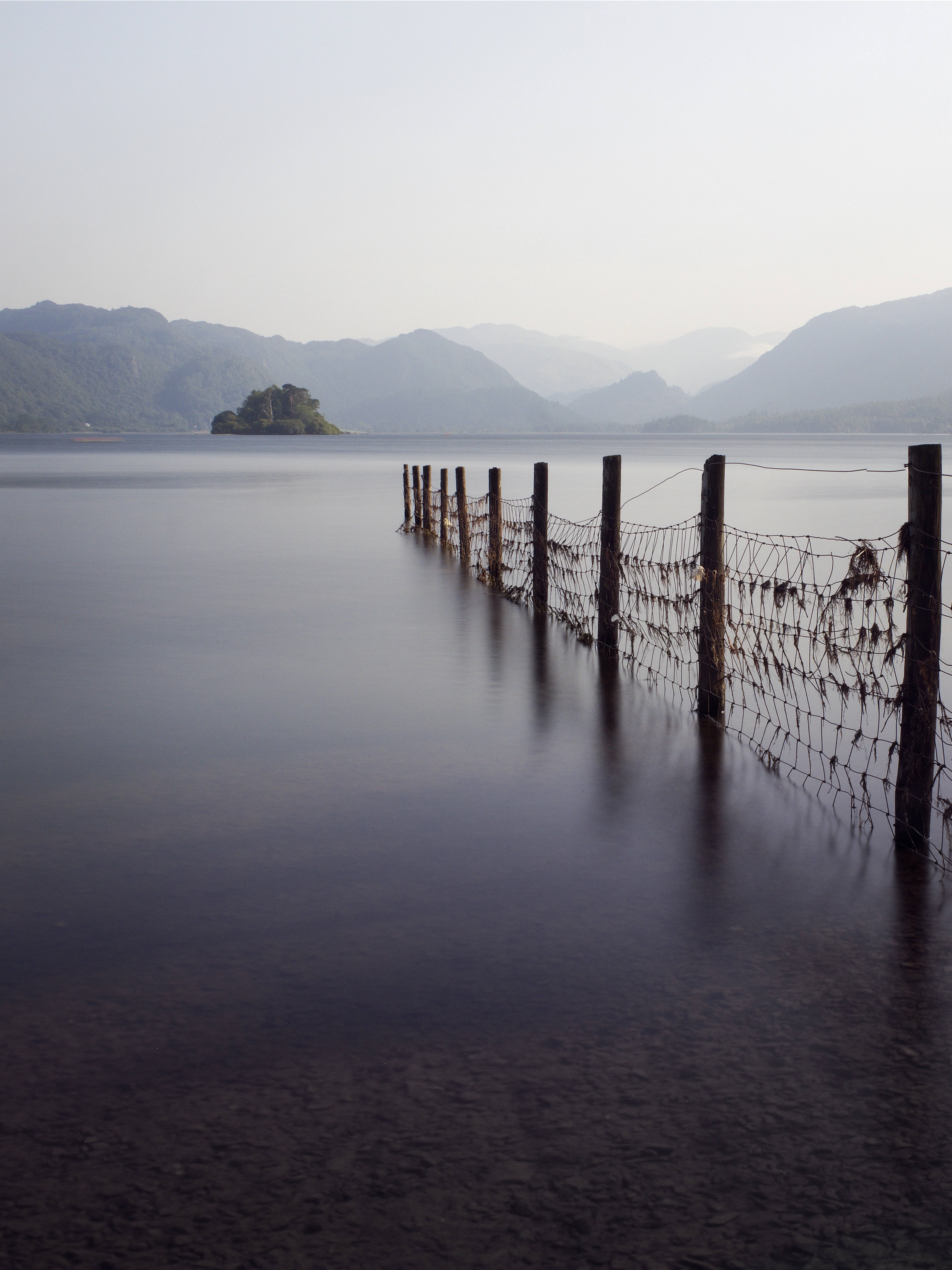 There's a strong chance this image contains the following subjects: fog,  water,  landscape,  lake,  sunset,  nature,  dawn,  beach,  pier. The photographer has provided the following description: This shot was taken close to Friar’s Crag by Derwentwater in Keswick. An old fence is seen leading the eye towards the distant mountains of the Lake District. Taken on a hot day in August 2025. A long exposure shot.  Photographer_name: Chris Michael. Title: Derwentwater, Lake Disrtrict. The photographer has provided the following description: This shot was taken close to Friar’s Crag by Derwentwater in Keswick. An old fence is seen leading the eye towards the distant mountains of the Lake District. Taken on a hot day in August 2025. A long exposure shot.  Make: fujifilm. Model: x-T5. Technical: 16-80mm lens at 16mm. ISO200. f16. 21 seconds. . . Published: 2025-08-24 15:50:08