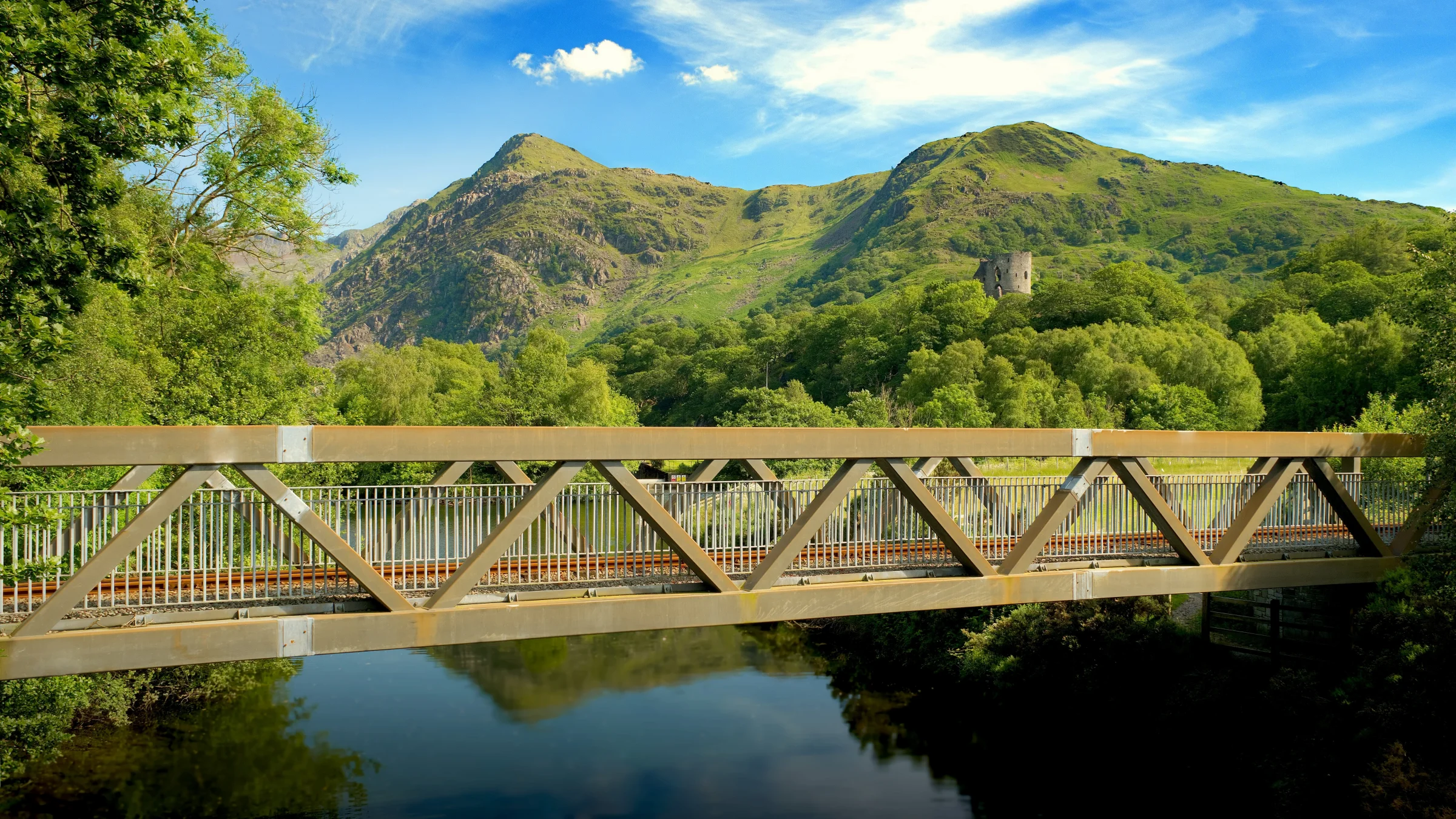 There's a strong chance this image contains the following subjects: tree,  sky,  water,  landscape,  lake,  travel,  nature,  wood,  river,  bridge,  wales,  snowdonia,  eryri,  eryrinationalpark,  nationalpark,  snowdon. The photographer has provided the following description: On a beautiful summer's day in Llanberis, photographing Snowdonia National Park. Dolbadarn Castle seen in the background. Photographer_name: Chris Michael. Title: Llanberis Bridge. The photographer has provided the following description: On a beautiful summer's day in Llanberis, photographing Snowdonia National Park. Dolbadarn Castle seen in the background. Make: fujifilm. Model: X-T5. Technical: 16-80mm. Published: 2025-09-09 14:21:32