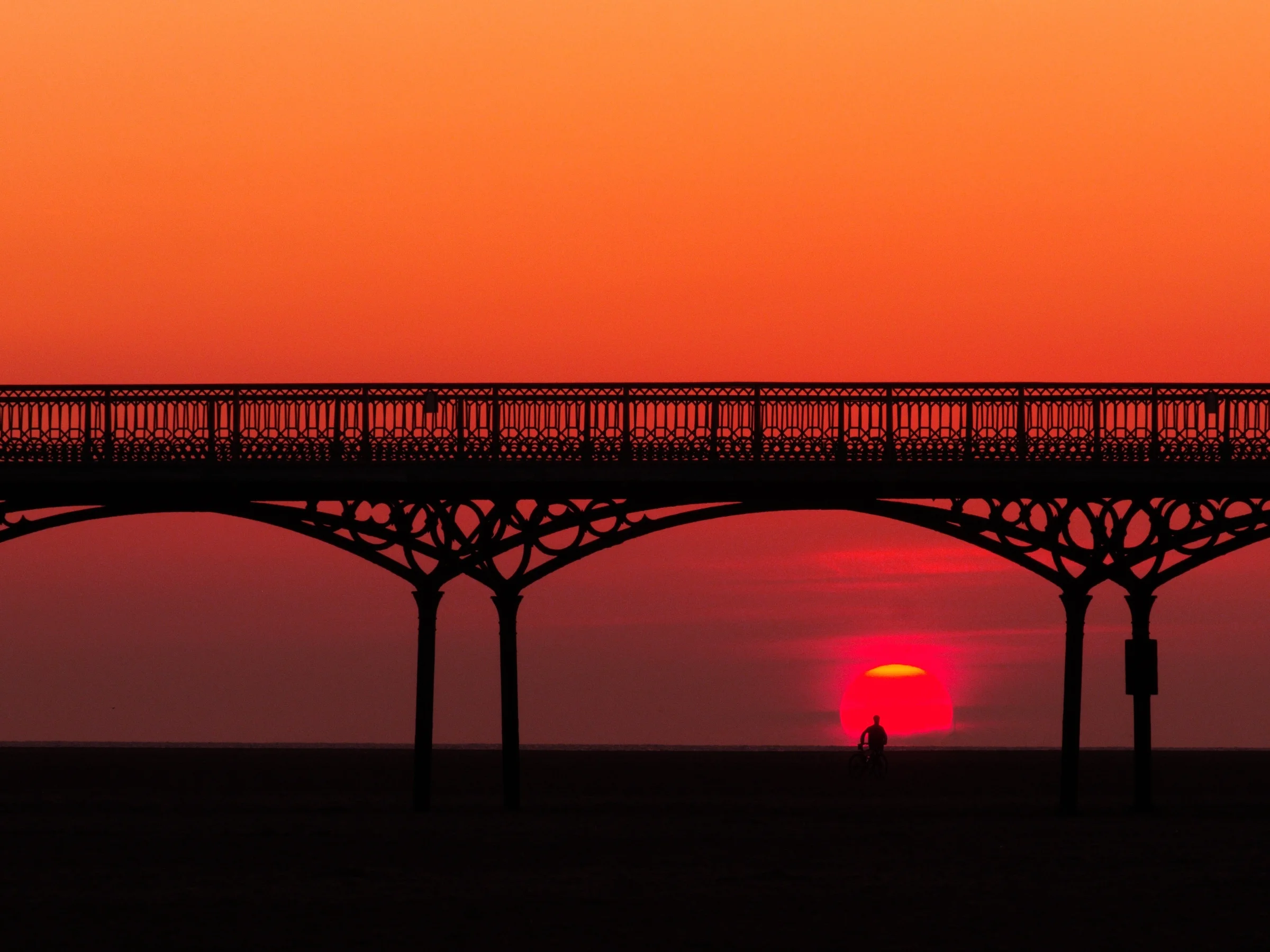 There's a strong chance this image contains the following subjects: water,  sea,  sunset,  dawn,  beach,  lancashire,  sun,  bridge,  ocean,  silhouette,  lytham,  lythamstannes,  pier. The photographer has provided the following description: Sat on St Annes Beach waiting for the sun to set under the pier and boom; not only did it glow bright red, but a cyclist just happened to push his bike into shot adding a gorgeous silhouette inside the sun. Photographer_name: Chris Michael. Title: Sunset at Lytham. The photographer has provided the following description: Sat on St Annes Beach waiting for the sun to set under the pier and boom; not only did it glow bright red, but a cyclist just happened to push his bike into shot adding a gorgeous silhouette inside the sun. Make: fujifilm. Model: X-T5. Technical: XF55-200mm lens at 200.. Published: 2025-09-07 17:03:42