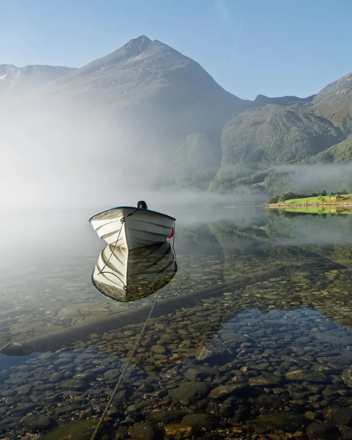 There's a strong chance this image contains the following subjects: water,  landscape,  lake,  mist. The photographer has provided the following description: In the Norwegian mountains, the misty lake mirrors the boat quiet reflection, surrounded by towering peaks. FOTO has this to say about the photograph: The photograph frames the boat like a quiet whisper on still water with beautifully sharp reflections. Its balanced composition gently guides the eye, inviting a moment of pause, a serene escape captured in subtle details and soft natural light. Source: https://www.instagram.com/ariel_a_k/ Photographer_name: Ariel Kozak. Title: Møre og Romsdal. The photographer has provided the following description: In the Norwegian mountains, the misty lake mirrors the boat quiet reflection, surrounded by towering peaks. FOTO has this to say about the photograph: The photograph frames the boat like a quiet whisper on still water with beautifully sharp reflections. Its balanced composition gently guides the eye, inviting a moment of pause, a serene escape captured in subtle details and soft natural light. Published: 2025-06-23 12:57:22