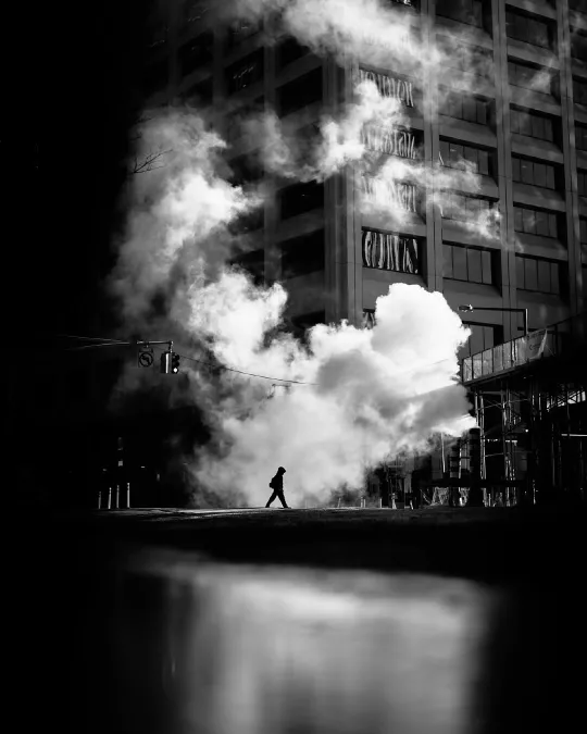 There's a strong chance this image contains the following subjects: street,  monochrome,  city,  dark,  smoke. The photographer has provided the following description: The experience of creating an image is more important than the final result FOTO has this to say about the photograph: This black and white image is anchored by a lone figure mid-stride, the composition is clean and deliberate, with contrast and shadow guiding the eye. The stillness of the space, paired with the solitary movement, creates a quiet, contemplative sense of calm. Source: https://www.instagram.com/philpenman/ Photographer_name: Philip Penman. Title: New York. The photographer has provided the following description: The experience of creating an image is more important than the final result FOTO has this to say about the photograph: This black and white image is anchored by a lone figure mid-stride, the composition is clean and deliberate, with contrast and shadow guiding the eye. The stillness of the space, paired with the solitary movement, creates a quiet, contemplative sense of calm. Published: 2025-06-27 13:34:33