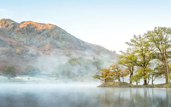 There's a strong chance this image contains the following subjects: tree,  sky,  fog,  water,  landscape,  mist,  fall,  nature. The photographer has provided the following description: Some lovely soft warm light catching the top of Loughrigg Fell with Misty reflections on Rydal Water Source: https://www.instagram.com/mark.worley.photography/ Photographer_name: Mark Worley. Title: Misty reflections. The photographer has provided the following description: Some lovely soft warm light catching the top of Loughrigg Fell with Misty reflections on Rydal Water Published: 2025-06-25 09:27:04