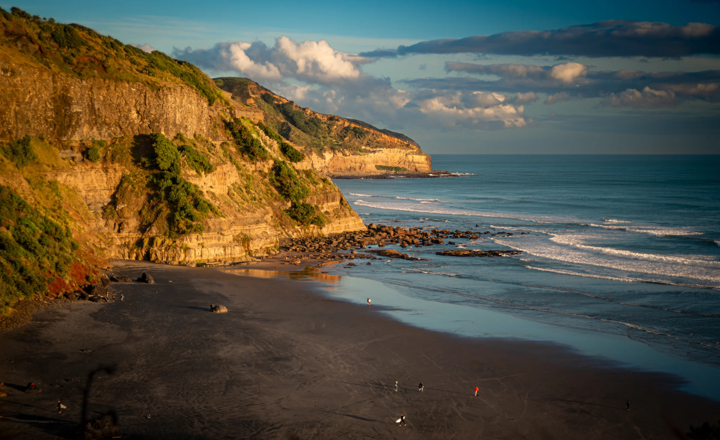There's a strong chance this image contains the following subjects: landscape,  sea,  beach,  cliff. The photographer has provided the following description: Muriwai Beach New Zealand Photographer_name: Robert A F van de Voort. Title: Black sand with surfers returning. The photographer has provided the following description: Muriwai Beach New Zealand Make: nikon. Model: D800. Technical: 210 - 70 mm Nikkor.. Published: 2026-06-03 04:16:14
